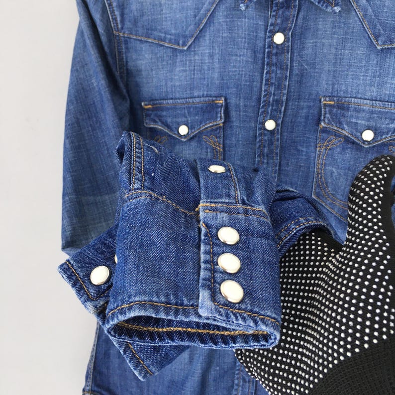 May include: A blue denim shirt with white buttons and two front pockets. The shirt has long sleeves with buttoned cuffs. The fabric is a medium wash denim.