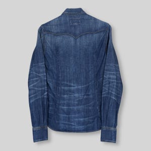 May include: A long-sleeved denim shirt in a medium blue wash. The shirt features a classic western-style yoke, buttoned cuffs, and a tailored fit. The back of the shirt is visible, showcasing the denim's texture and subtle fading.