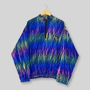 Puede incluir: Chaqueta estampada azul, verde y rosa con cierre de media cremallera. La chaqueta tiene cuello alto y un pequeño parche con el logo en el pecho. La chaqueta está colgada en una percha de madera.