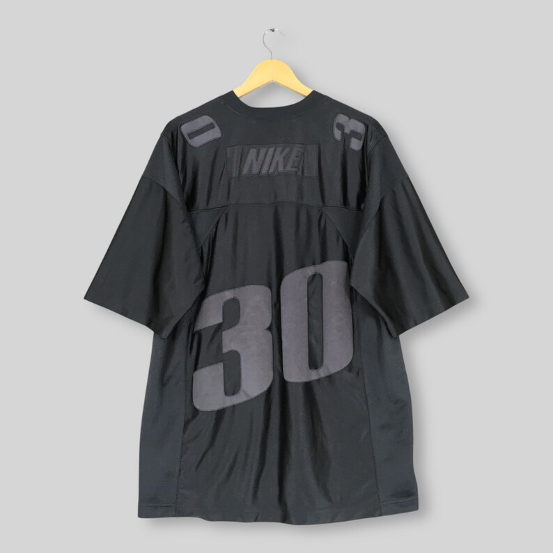 Puede incluir: Camiseta negra de Nike con el n&uacute;mero 30 en grandes numerales grises en la parte delantera y trasera. El logotipo de Nike est&aacute; centrado en el pecho. La camiseta est&aacute; colgada en una percha de madera.
