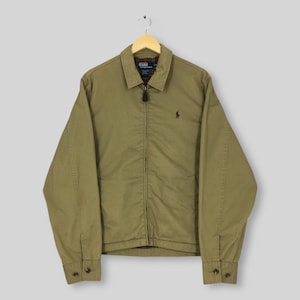 May include: A khaki-colored zip-up jacket with a collared neckline and long sleeves. The jacket features a small embroidered logo on the chest and buttoned cuffs. The jacket is hanging on a wooden hanger.
