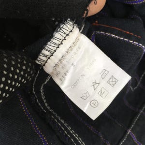 以下が含まれることがあります： NO. 561-0815、SIZE T、COTTON 100%と書かれた黒い衣類ケアラベル。白い長方形のタグには、洗濯、漂白、アイロン掛け、ドライクリーニングの標準的なケアシンボルが記載されており、紫色のステッチで暗い色の生地に縫い付けられています。