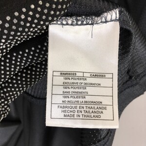 Puede incluir: Primer plano de una etiqueta blanca con texto negro, adherida a una prenda gris oscuro. La etiqueta incluye detalles como "RN#56323", "100% POLYESTER" y "MADE IN THAILAND". La prenda tiene un patr&oacute;n de puntos.