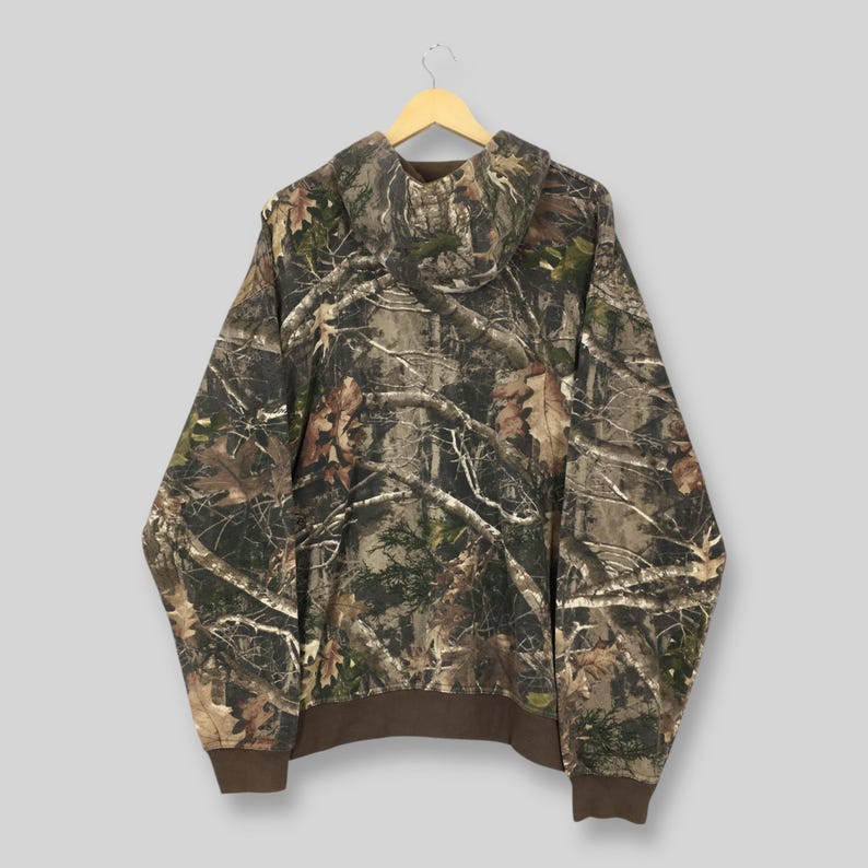 May include: A camouflage hooded sweatshirt with a brown hem and cuffs. The hoodie features a pattern of brown, green, and tan branches and leaves. The sweatshirt is suspended from a wooden hanger.