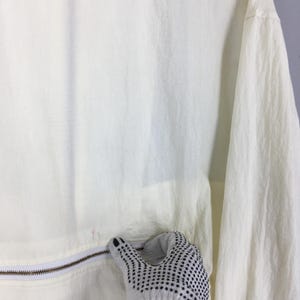 May include: A lightweight, off-white windbreaker jacket featuring a crinkled texture, long sleeves, and a horizontal zipper closure across the midsection. The fabric has a semi-sheer quality, and the garment includes a white zipper track.