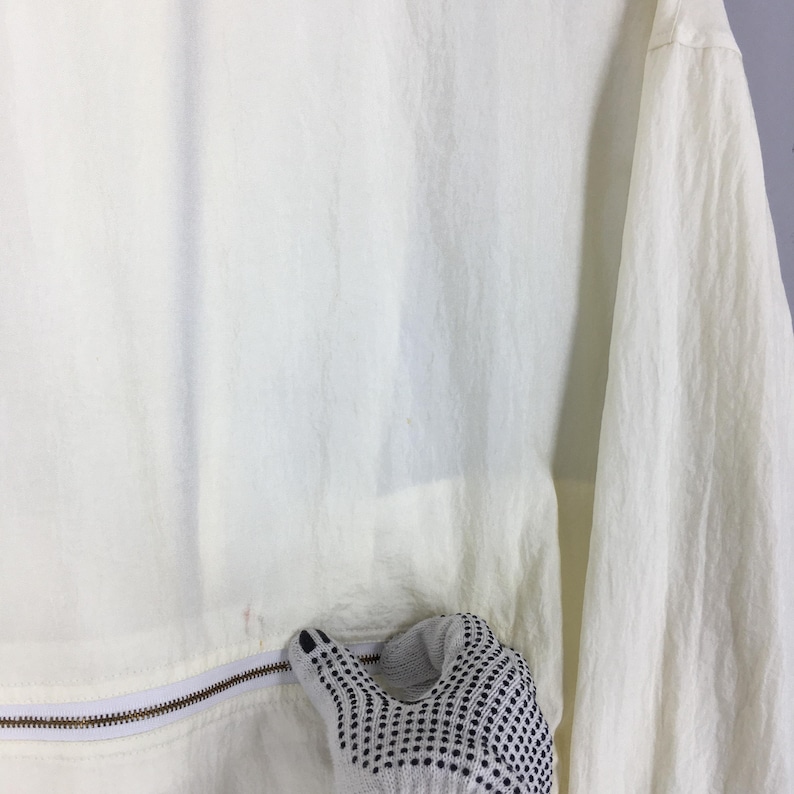 May include: A lightweight, off-white windbreaker jacket featuring a crinkled texture, long sleeves, and a horizontal zipper closure across the midsection. The fabric has a semi-sheer quality, and the garment includes a white zipper track.