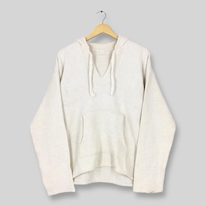 Può includere: Una felpa con cappuccio color crema con tasca a marsupio e scollatura con coulisse. Il top a maniche lunghe è realizzato in tessuto strutturato e appeso a una gruccia di legno su sfondo grigio chiaro. La felpa ha una vestibilità ampia.