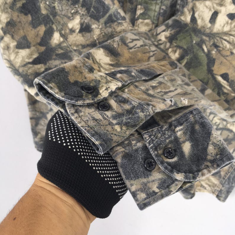 May include: Close-up of a camouflage button-down shirt. The shirt features a pattern of brown, green, and tan, with black buttons on the cuffs. The fabric appears to be a soft, brushed material.