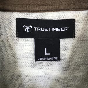 May include: Close-up of a clothing label. The black label displays the brand name "TRUETIMBER" in white. Below, a smaller black label shows the letter "L" and the text "MADE IN PAKISTAN". The sweatshirt fabric is light grey with a brown collar.