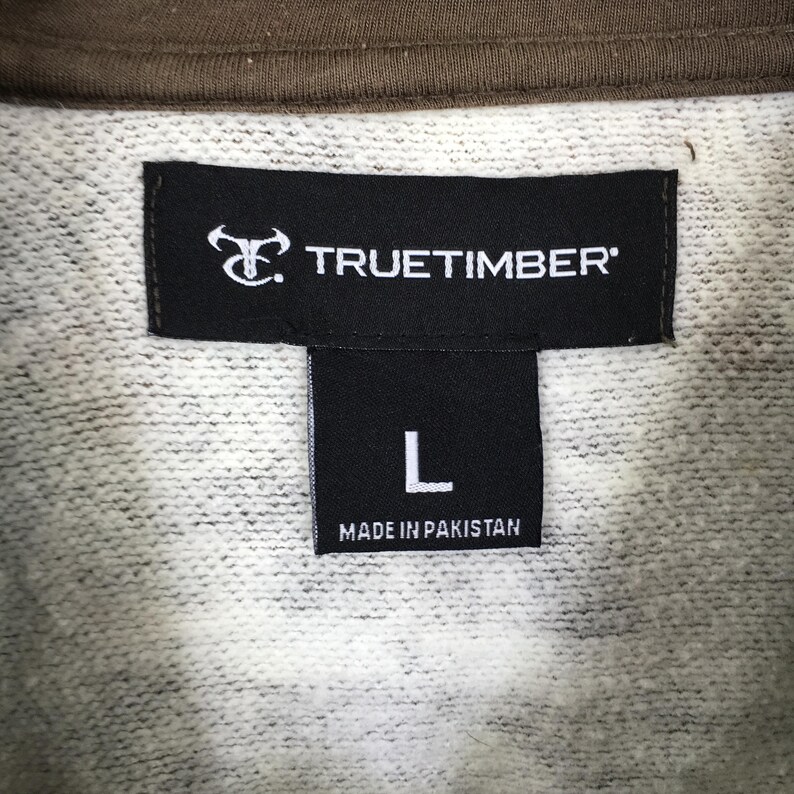 May include: Close-up of a clothing label. The black label displays the brand name "TRUETIMBER" in white. Below, a smaller black label shows the letter "L" and the text "MADE IN PAKISTAN". The sweatshirt fabric is light grey with a brown collar.