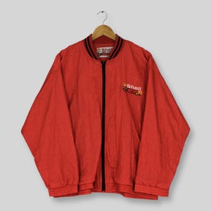 May include: A red zip-up bomber jacket featuring a black front zipper and a ribbed collar with black stripes. The left chest displays an embroidered Shell F1 logo with a race car graphic. The interior neck label also features the Shell F1 branding.
