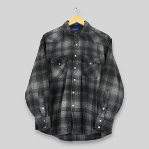 Pode incluir: Uma camisa de manga comprida com botões num padrão de xadrez cinzento e preto. A camisa apresenta um colarinho pontiagudo, dois bolsos no peito com abas e botões de pressão prateados na frente, nos punhos e nas abas dos bolsos. A etiqueta Pendleton é visível no interior do colarinho.