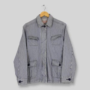 Può includere: Una giacca a maniche lunghe con un motivo a righe verticali blu navy e bianco. La giacca presenta un colletto a punta, chiusura frontale con bottoni, due tasche sul petto con patta e due tasche inferiori con patta, tutte chiuse da bottoni scuri.
