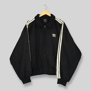 Könnte beinhalten: Schwarze Adidas-Trainingsjacke mit weißen Streifen an den Ärmeln und dem Adidas-Logo. Die Jacke hat einen durchgehenden Reißverschluss, einen Kragen sowie gerippte Bündchen und Saum. Sie hängt an einem Holzkleiderbügel.