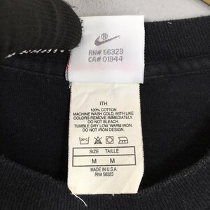 Puede incluir: Primer plano de una etiqueta de camiseta negra con el logo de Nike y texto. La etiqueta incluye instrucciones de lavado, informaci&oacute;n de talla (M) y el texto "MADE IN U.S.A. RN# 56323".