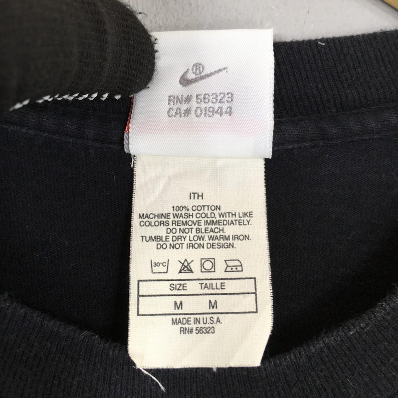 Puede incluir: Primer plano de una etiqueta de camiseta negra con el logo de Nike y texto. La etiqueta incluye instrucciones de lavado, informaci&oacute;n de talla (M) y el texto "MADE IN U.S.A. RN# 56323".