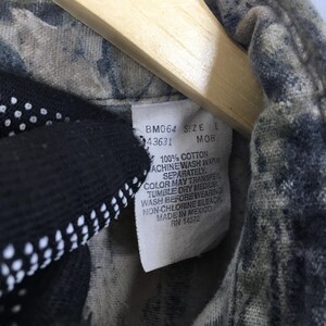 May include: Close-up of a camouflage-patterned shirt with a tag. The tag reads "BM064 SIZE L 43631 MOB 100% COTTON MACHINE WASH WARM SEPARATELY. COLOR MAY TRANSFER TUMBLE DRY MEDIUM WASH BEFORE WEARING NON-CHLORINE BLEACH MADE IN MEXICO RN 14372."