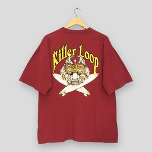 May include: A dark red short-sleeve t-shirt featuring a graphic on the back. The design includes the yellow text Killer Loop arched over a detailed illustration of a traditional Balinese mask with two crossed white blades positioned underneath.