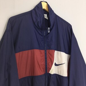 Pu&ograve; includere: Giacca a vento blu navy e rossa con un pannello bianco e un logo Nike nero. La giacca ha una cerniera completa e un colletto alto. La giacca &egrave; appesa a una gruccia di legno.