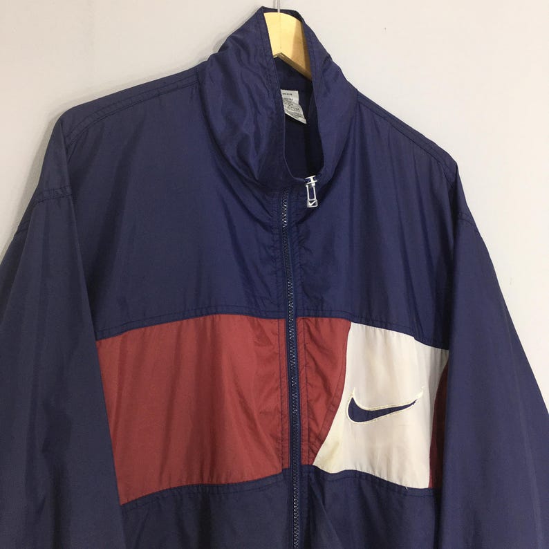 Pu&ograve; includere: Giacca a vento blu navy e rossa con un pannello bianco e un logo Nike nero. La giacca ha una cerniera completa e un colletto alto. La giacca &egrave; appesa a una gruccia di legno.