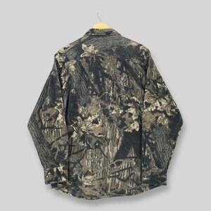 May include: A long-sleeved button-down shirt with a camouflage pattern. The shirt features a blend of green, brown, and gray tones, mimicking a forest environment. The shirt is hanging on a wooden hanger.