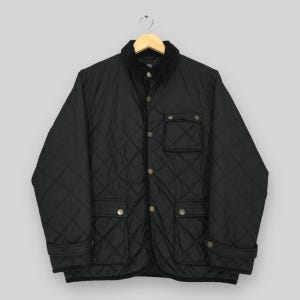 Puede incluir: Chaqueta acolchada negra con cuello de pana y botones dorados. La chaqueta tiene dos bolsillos delanteros y dos bolsillos en el pecho. La chaqueta está colgada de una percha de madera.