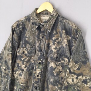 May include: A long-sleeved button-down shirt with a camouflage pattern in shades of green, brown, and gray. The shirt has two chest pockets and a collar. The tag reads "NORTHWESTERN".