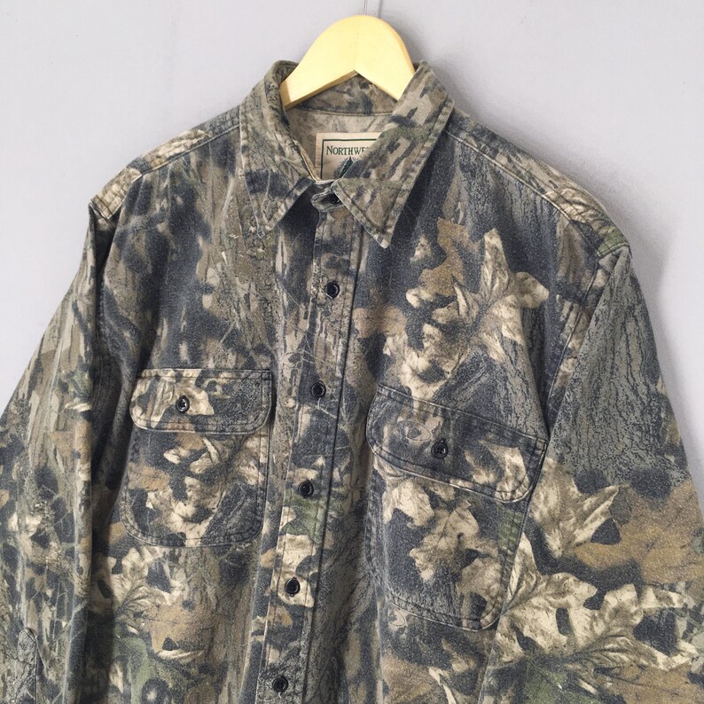 May include: A long-sleeved button-down shirt with a camouflage pattern in shades of green, brown, and gray. The shirt has two chest pockets and a collar. The tag reads "NORTHWESTERN".
