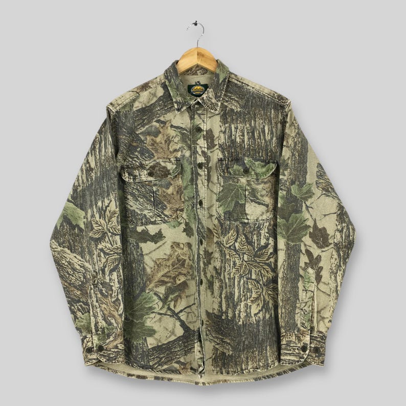 Puede incluir: Una camisa de manga larga con estampado de camuflaje. La camisa presenta un dise&ntilde;o de bosque con tonos verdes, marrones y beige. Tiene dos bolsillos en el pecho y est&aacute; colgada de una percha de madera.