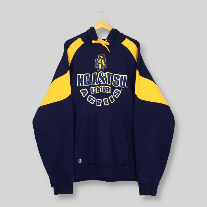May include: Navy blue hoodie with yellow shoulder and hood panels. The front displays the text "NC A&T SU EST. 1891 AGGIES" in yellow and white. The hoodie has a front pocket and a drawstring hood.