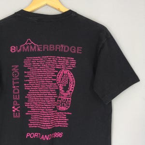 Puede incluir: Camiseta negra con texto y gr&aacute;ficos rosas. El texto dice "SUMMERBRIDGE EXPEDITION" con una lista de nombres y "PORTLAND 1996". Tambi&eacute;n se ve un gr&aacute;fico de huella de bota rosa.