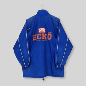 May include: A royal blue windbreaker jacket featuring a high collar and long sleeves with white piping accents. The back displays a red rectangular logo with a white rhinoceros icon above the orange text ECKO and smaller text below.