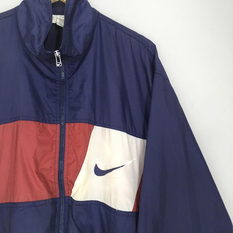 Pu&ograve; includere: Giacca a vento Nike vintage blu navy, rossa e bianca. La giacca presenta una cerniera completa, un colletto alto e il logo Nike swoosh sul davanti. Il design a blocchi di colore include una striscia rossa sul petto.