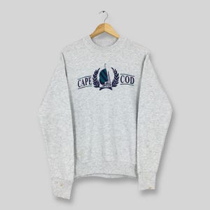 Op de afbeelding: Een lichtgrijs sweatshirt met de tekst "CAPE COD" boven en onder een zeilbootafbeelding in donkerblauw en groen. De sweatshirt heeft een ronde hals en lange mouwen.