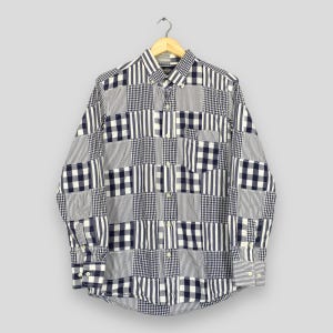 May include: A long-sleeved shirt with a patchwork design in navy blue and white. The shirt features a mix of patterns, including stripes and checks. It is displayed on a wooden hanger against a plain background.