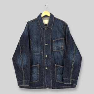 May include: A dark blue denim jacket with four front pockets and button closures. The jacket has a collared neckline and is hanging on a wooden hanger. The stitching is a contrasting light brown color.