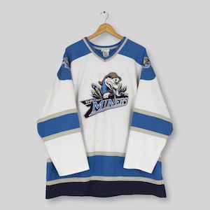 May include: White and blue hockey jersey featuring the text "Grumpy Miners" and a cartoon miner graphic. The jersey has blue and grey stripes on the sleeves and hem. It is hanging on a wooden hanger.