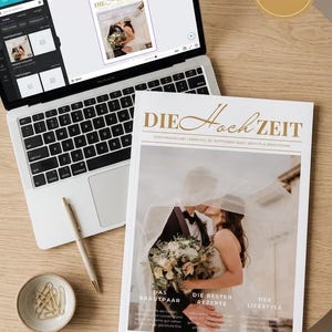May include: A laptop displaying a design interface for a wedding magazine titled DIE HOCHZEIT, alongside a printed copy of the magazine featuring a couple under a veil, a gold pen, and a small bowl of gold paper clips on a round wooden surface.