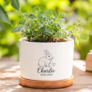 May include: A white ceramic cylindrical planter featuring a black line drawing of a rabbit, a small heart, and the text "Charlie 2006-2024". The planter sits on a circular light brown bamboo saucer and contains a small green leafy plant.