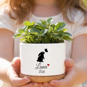 Può includere: Un vaso cilindrico in ceramica bianca con base in bambù contenente una pianta verde. La parte anteriore presenta una silhouette nera di un coniglio e una farfalla, il nome "Luna" in corsivo nero, un piccolo cuore rosso e l'anno "2026".