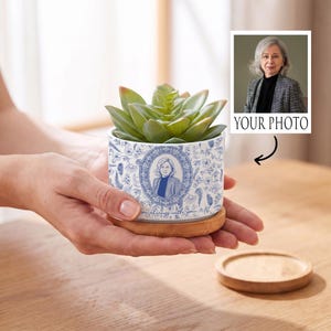 Może przedstawiać: Biała ceramiczna doniczka z niebieskim wzorem kwiatowym i centralną owalną ramką zawierającą portret osoby. Doniczka stoi na okrągłej bambusowej podstawie i zawiera zielony sukulent. Tekst "I am always with you" jest nadrukowany pod portretem.