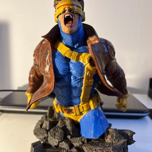 Może przedstawiać: Szczegółowe popiersie postaci Cyclops z X-Men, w niebiesko-żółtym kostiumie z brązową kurtką. Postać ma otwarte usta i żółty wizjer. Podstawa to skalisty teren z logo X-Men.