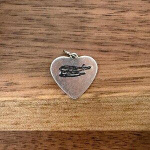 May include: A silver heart-shaped pendant with a black airplane design. The pendant is suspended from a small loop and is set against a wooden background. The airplane is a seaplane.