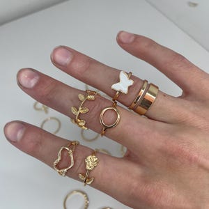 Peut inclure: Ensemble de bagues dorées avec divers motifs : papillon, cœur, rose et feuilles. Les bagues sont présentées sur une main, sur fond blanc. Les bagues sont faites d'un matériau métallique brillant.