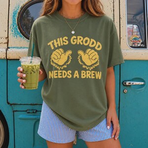 May include: An olive green short-sleeve t-shirt featuring yellow graphic text that reads THIS GRODD NEEDS A BREW, accompanied by two yellow illustrations of furry hands with thumbs pointing inward, paired with blue and white vertical striped shorts.