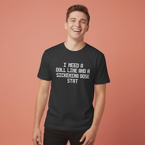 以下が含まれることがあります： 「I NEED A DOLL LINE AND A SICKENING DOSE STAT」と白い文字で書かれた黒いTシャツ。クルーネックのシャツは柔らかい素材で作られています。Tシャツを着ている人は笑顔です。背景は単色のピーチ色です。