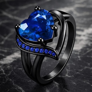 May include: A black ring featuring a heart-shaped blue gemstone. The ring has a double band design and is embellished with smaller blue gemstones along a curved band. The ring is set against a dark background, with a focus on the jewellery.