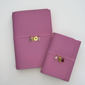 May include: Two pink leather journals of different sizes. Each journal has a pink elastic band with gold-coloured charms. The larger journal has a charm with a green stone and a gold-coloured sun. The smaller journal has a charm with a white stone and a red stone.