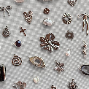 Peut inclure: Une collection de breloques et pendentifs en métal assortis de formes variées, incluant des cœurs, fleurs, nœuds, soleils, croix et tranches de pierre naturelle aux bords métalliques, disposés sur une surface texturée gris clair.