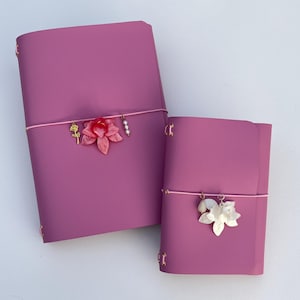 May include: Two magenta journals with rounded corners, each secured by a thin pink elastic band. The larger journal features a gold rose charm, a pink flower charm, and a pearl charm. The smaller journal features a white flower and a teardrop charm.